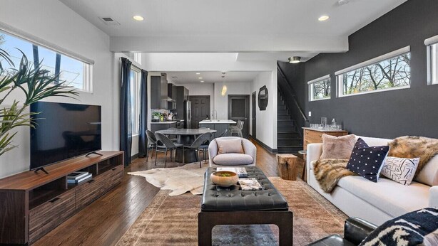 A modern living room with a TV, sofa, and coffee table. There is a dining area with a table and chairs. The room has large windows and a staircase. - Immaculate Meets Comfort! 3 BR Spacious Home! (Nashville)