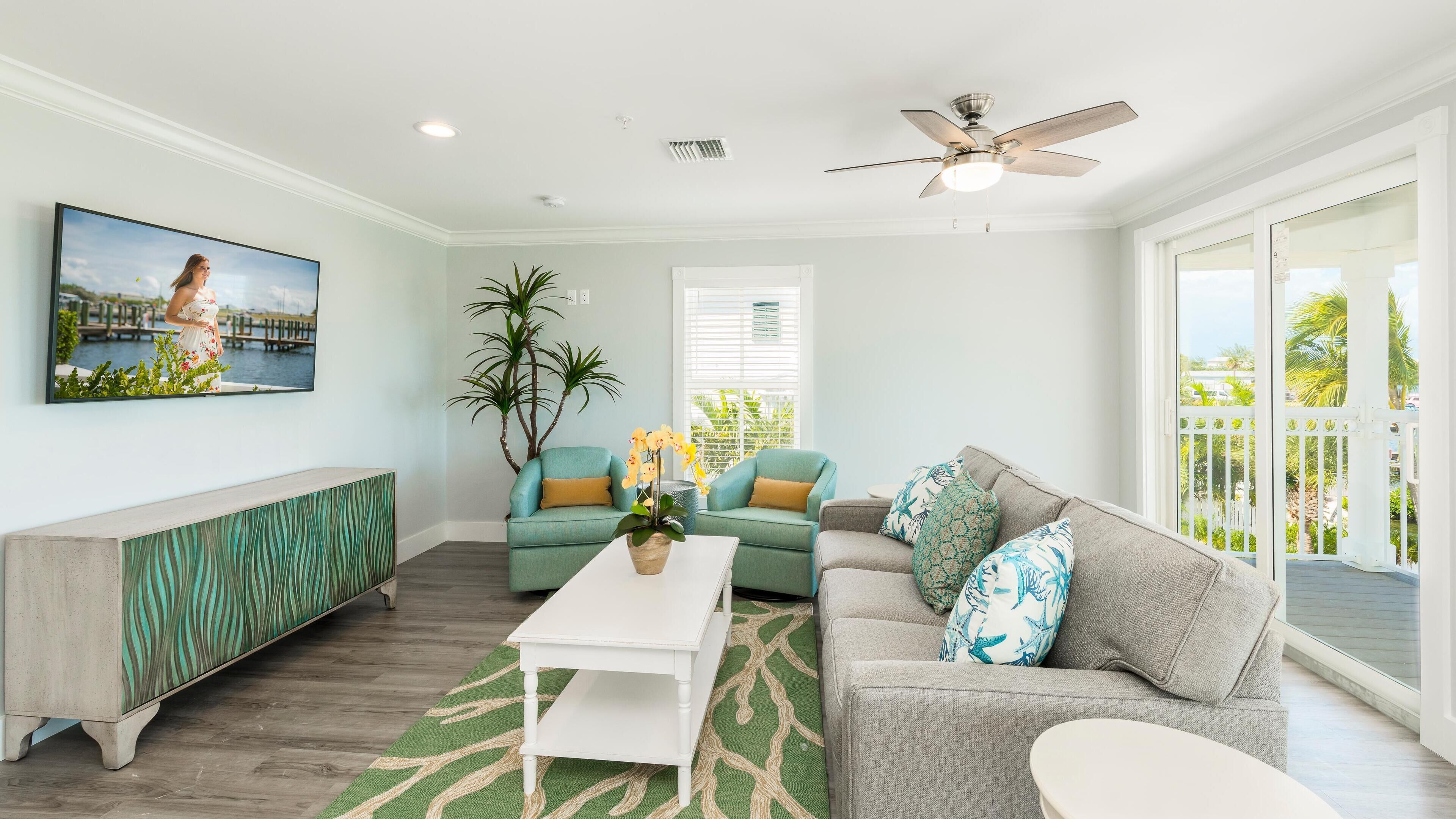 A living room with a TV, teal and gray sofas, a white coffee table, and a ceiling fan.