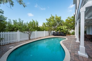 Una piscina con valla blanca, rodeada de árboles y un patio con sillas y una mesa.