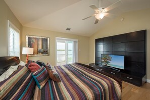 Un dormitorio con una cama grande, un ventilador de techo, un televisor sobre un mueble negro y un ventanal corredizo hacia el exterior.