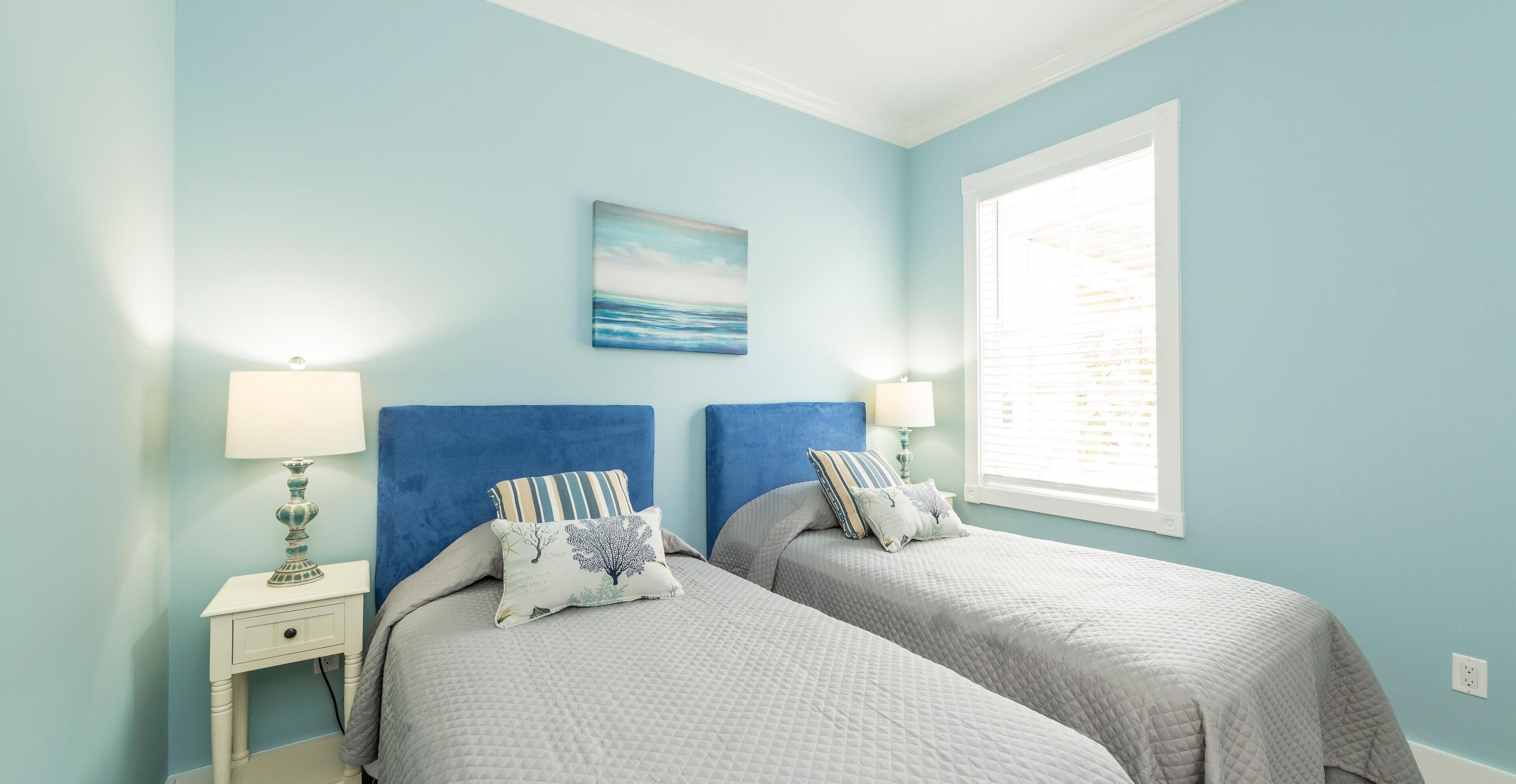 A bedroom with two beds, a nightstand with a lamp, a painting on the wall, and a window with blinds.