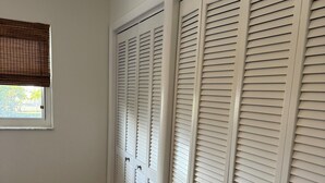 Une pièce avec un plancher en bois, des murs blancs et une fenêtre avec des stores. On y trouve des portes blanches dont le sommet est ajouré.