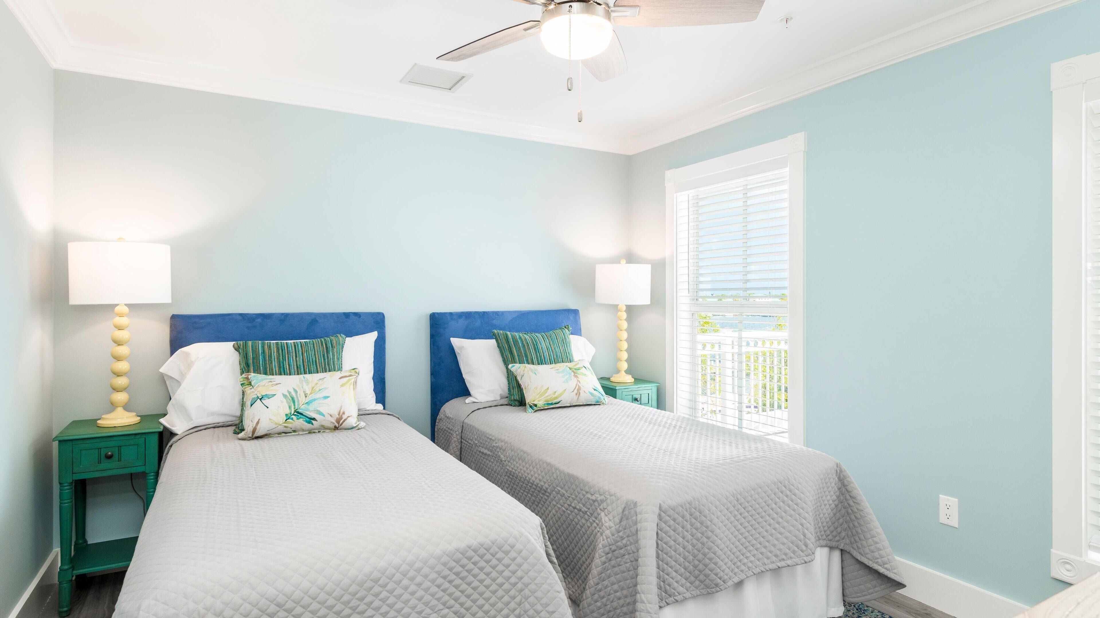 A bedroom with two beds, each with a blue headboard, white bedding, and decorative pillows. There are bedside tables with lamps and a ceiling fan.