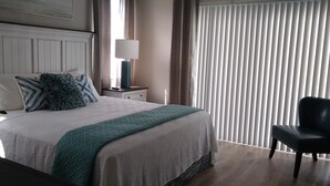 Een opgemaakt bed met een wit-groene sprei, een bedlampje en een stoel in een kamer met een groot raam en jaloezieën.