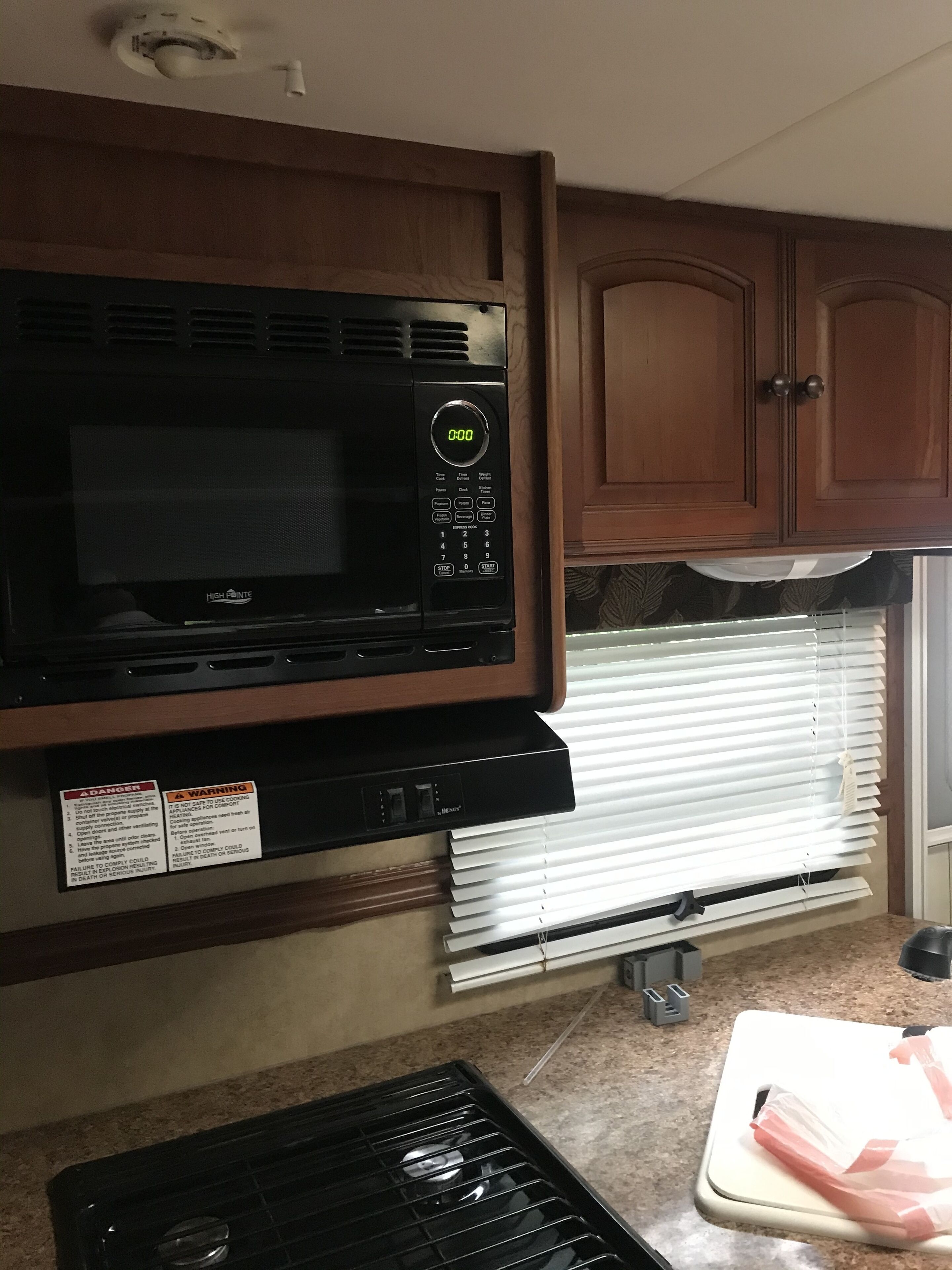 A kitchen with a microwave, wooden cabinets, a window with blinds, and a countertop with a cutting board and plastic bags.