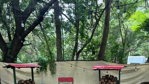 Une terrasse en bois mène à un espace détente avec deux tables à toit rouge et un foyer, le tout entouré d’arbres et d’une zone abritée où sont empilés des bûches.