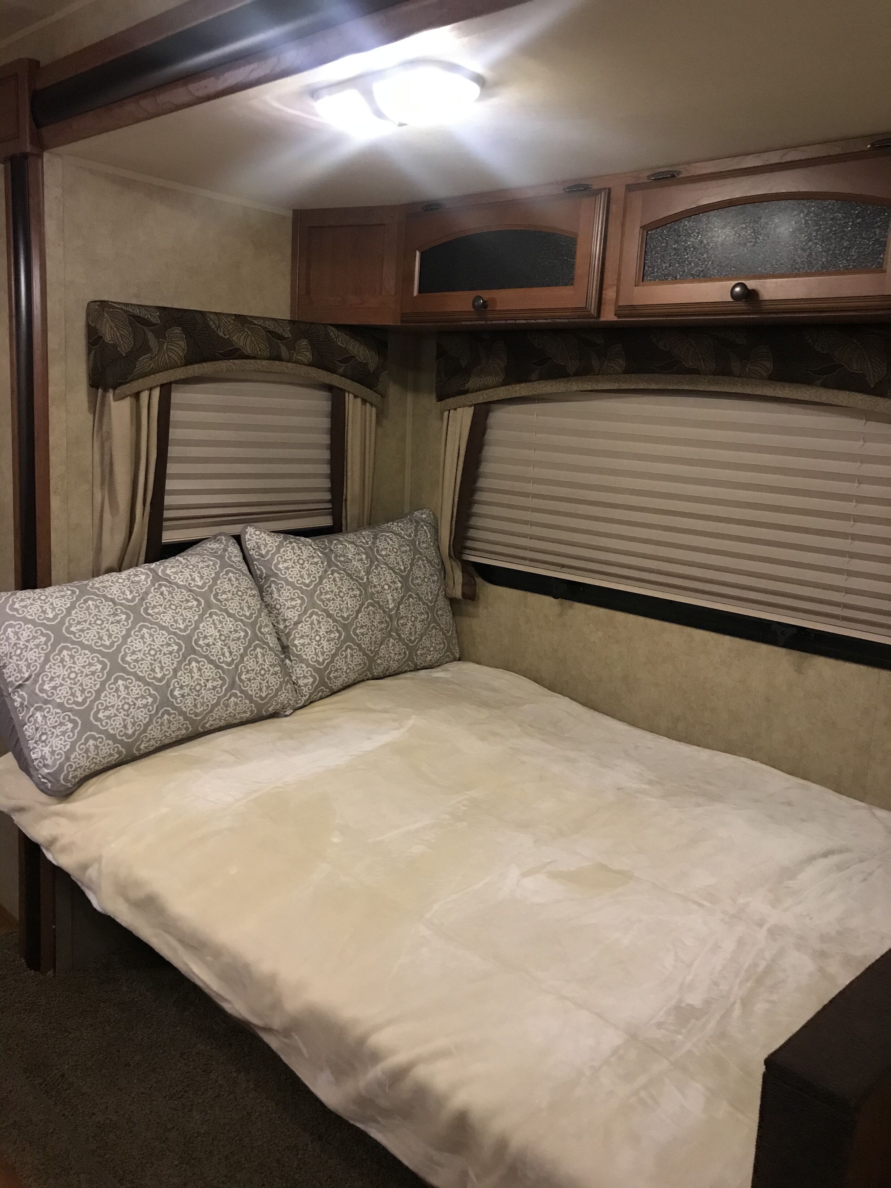Interior compacto de una caravana con una cama, dos ventanas con persianas y una luz en el techo.