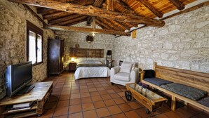 Una habitación rústica con paredes de piedra, techo de madera, cama, sofá, televisor sobre una mesa de madera y una pequeña mesa con lámpara.