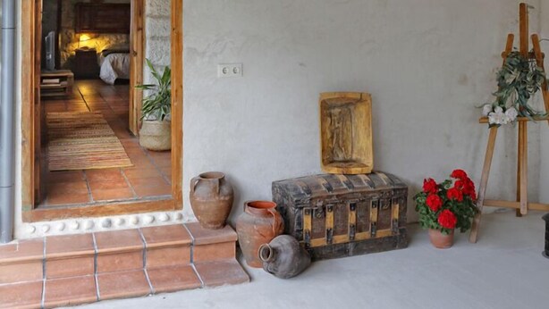 Un interior rústico con techo de madera, un vano que da a otra habitación, una pequeña mesa con un florero y un cuadro en la pared.