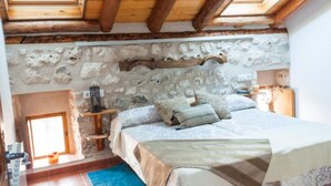 Un dormitorio con pared de piedra, vigas de madera, una cama con almohadas, una mesita de noche y una pequeña mesa con lámpara.