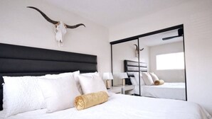 Una habitación de hotel con una cama grande, ropa de cama blanca, un cabecero negro, una mesita de noche con una lámpara y un espejo grande que refleja la habitación.