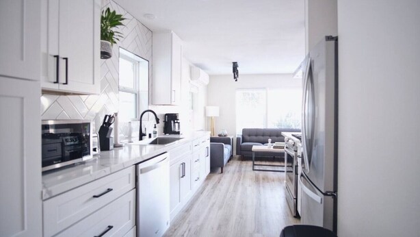 Una cocina moderna con armarios blancos, horno integrado y cafetera. En la sala de estar hay un sofá gris y una mesita de centro.