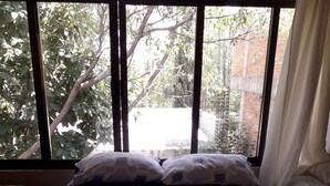 Une chambre à coucher avec un lit recouvert d’une courtepointe à motifs floraux bleus et blancs, deux oreillers et une fenêtre donnant sur des arbres et un mur de briques.