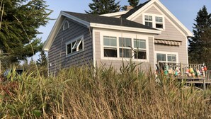 A two-story house with a grey exterior, white trim, and a porch with colorful decorations. - Cottage on Sawyer’s Cove (Jonesport)