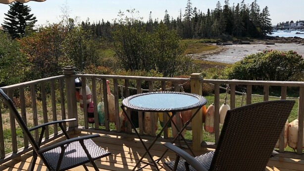 A covered outdoor area with a round table and two chairs, overlooking a beach and forested area. - Cottage on Sawyer’s Cove (Jonesport)
