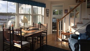 A dining area with a table and chairs, a lamp, and a view of the sea through large windows. - Cottage on Sawyer’s Cove (Jonesport)