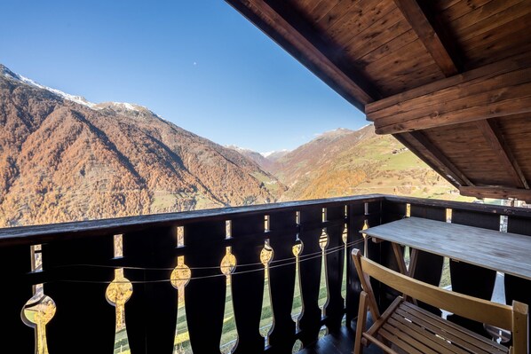 A wooden balcony with a table and chairs, overlooking a mountainous landscape with snow-capped peaks.
