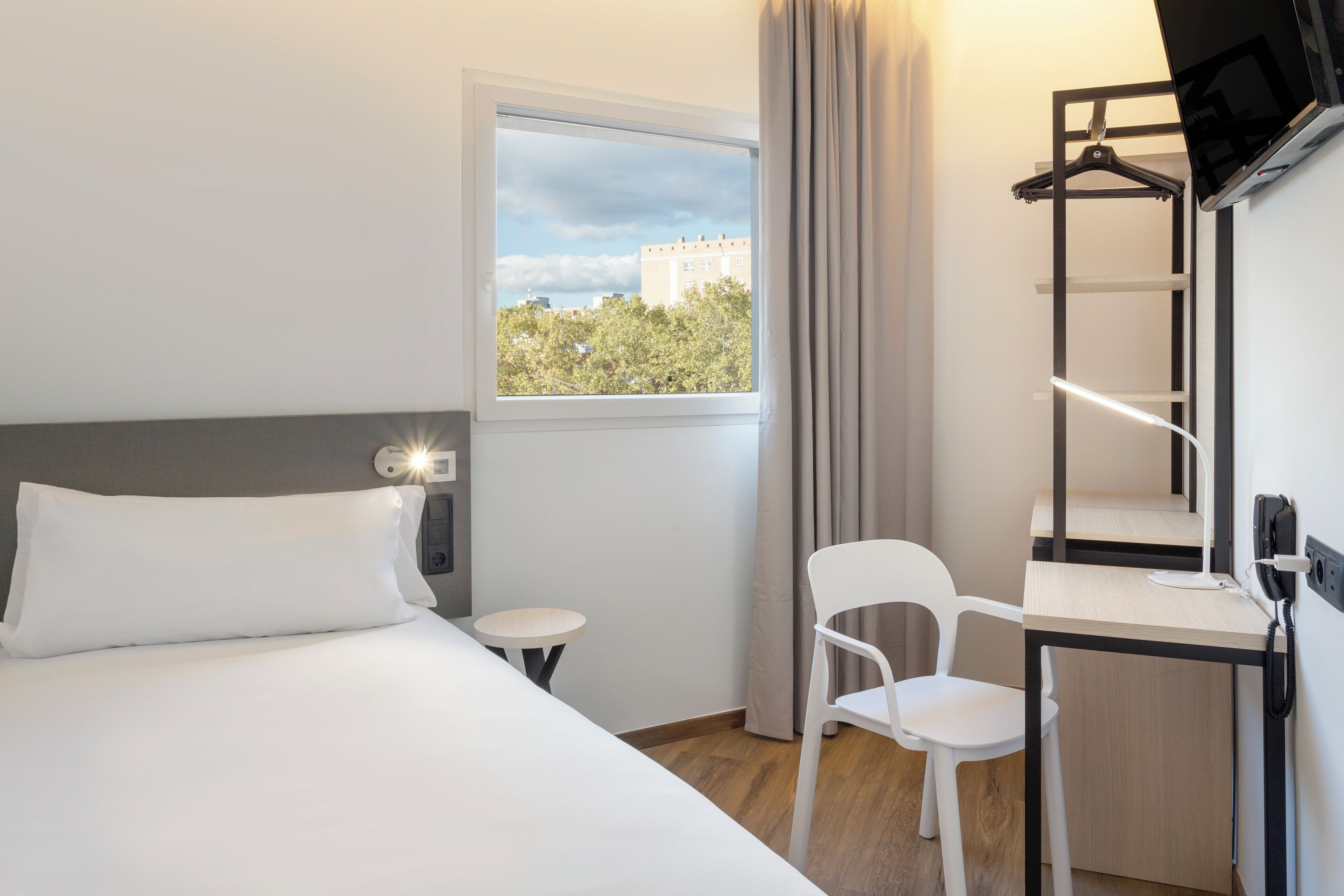 A hotel room with a bed, a desk with a chair, a small table, a window with a view of trees and buildings, and a TV mounted on the wall.