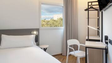 A hotel room with a bed, a desk with a chair, a small table, a window with a view of trees and buildings, and a TV mounted on the wall.