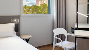 Habitación de hotel con cama, una pequeña mesa redonda, una silla blanca, un escritorio con una computadora y una ventana con vistas a edificios y árboles.
