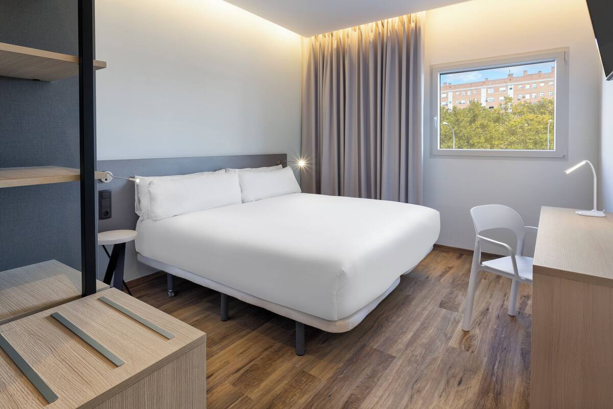 A modern hotel room with a large bed, a desk, and a chair. There is a window with a view of buildings and trees.