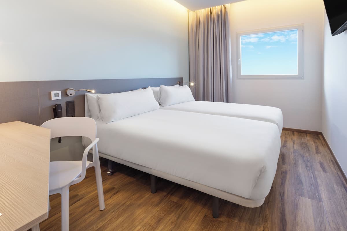 A hotel room with two beds, a desk, a chair, a window with curtains, and a wall-mounted lamp.