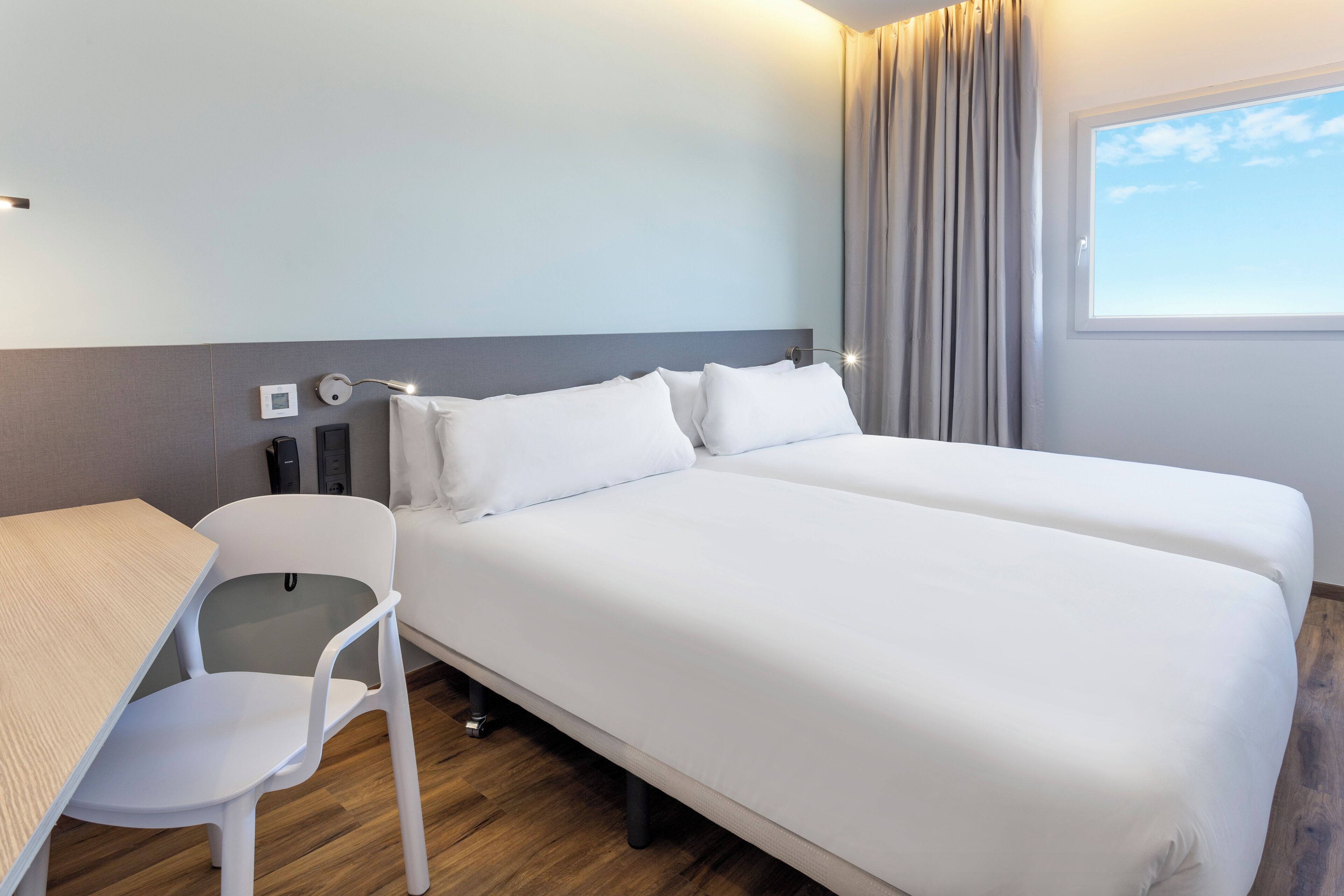 A hotel room with two beds, a desk, a chair, a window with curtains, and a thermostat on the wall.