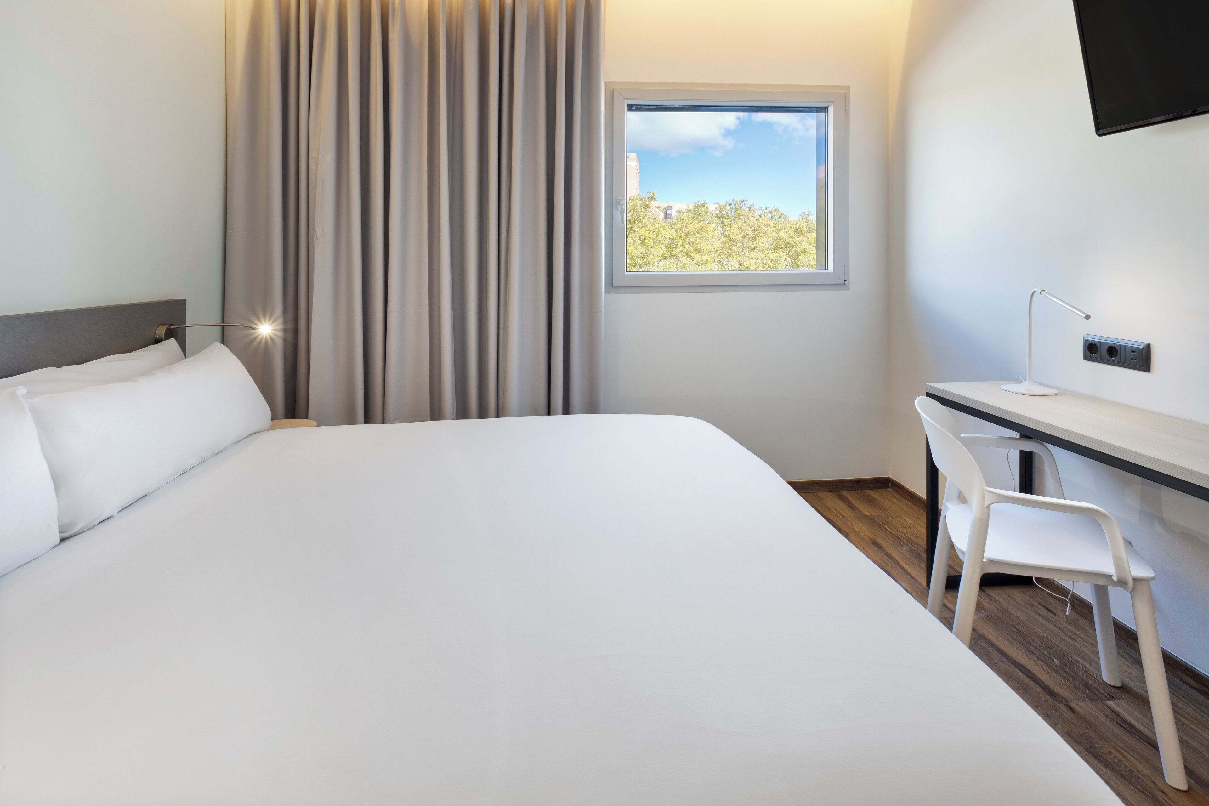 A hotel room with a large bed, a desk with a chair, a window with a view of trees, and a mounted TV.