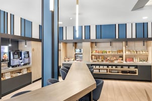 Um café moderno com uma mesa de madeira comprida, assentos e diversas opções de comida e bebida expostas em prateleiras.