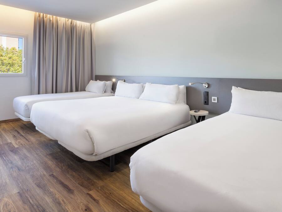 A hotel room with three single beds, a small bedside table, a window with curtains, and a wooden floor.