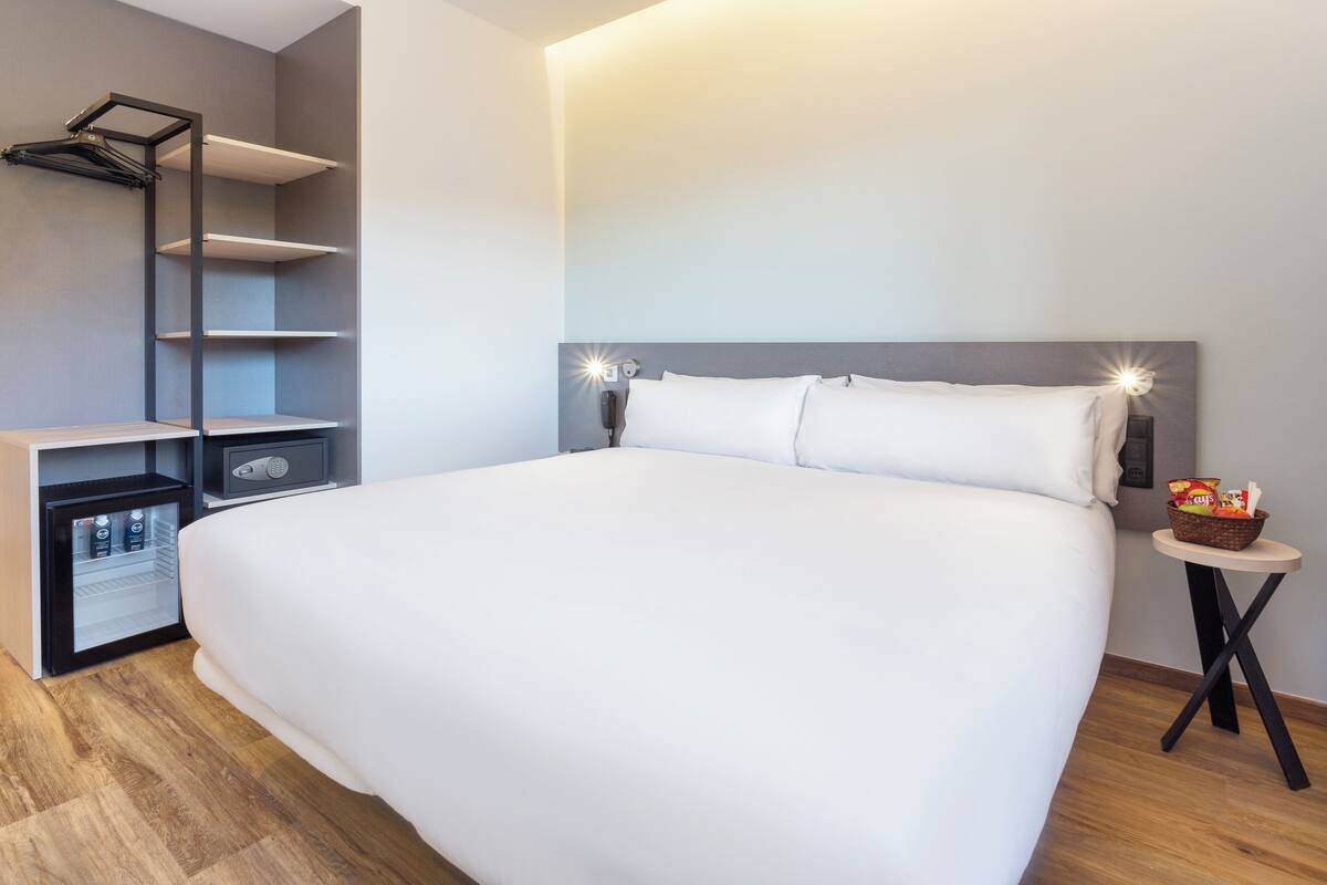 A modern hotel room with a large bed, built-in shelving, a minibar, and a small table with a basket of snacks.