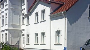 Ein Gebäude mit weißer Fassade, rotem Dach und einem gepflasterten Eingangsbereich, in dessen Nähe ein Auto geparkt ist.