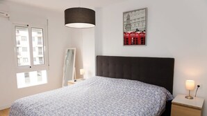 Un dormitorio con una cama, una mesita de noche con lámpara, una ventana y una obra de arte colgada en la pared.