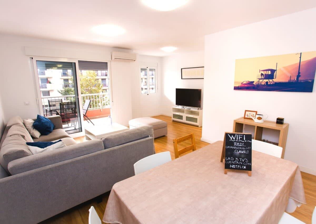 A modern living room with a sofa, a dining table, and a TV. There is a sign indicating WiFi availability.