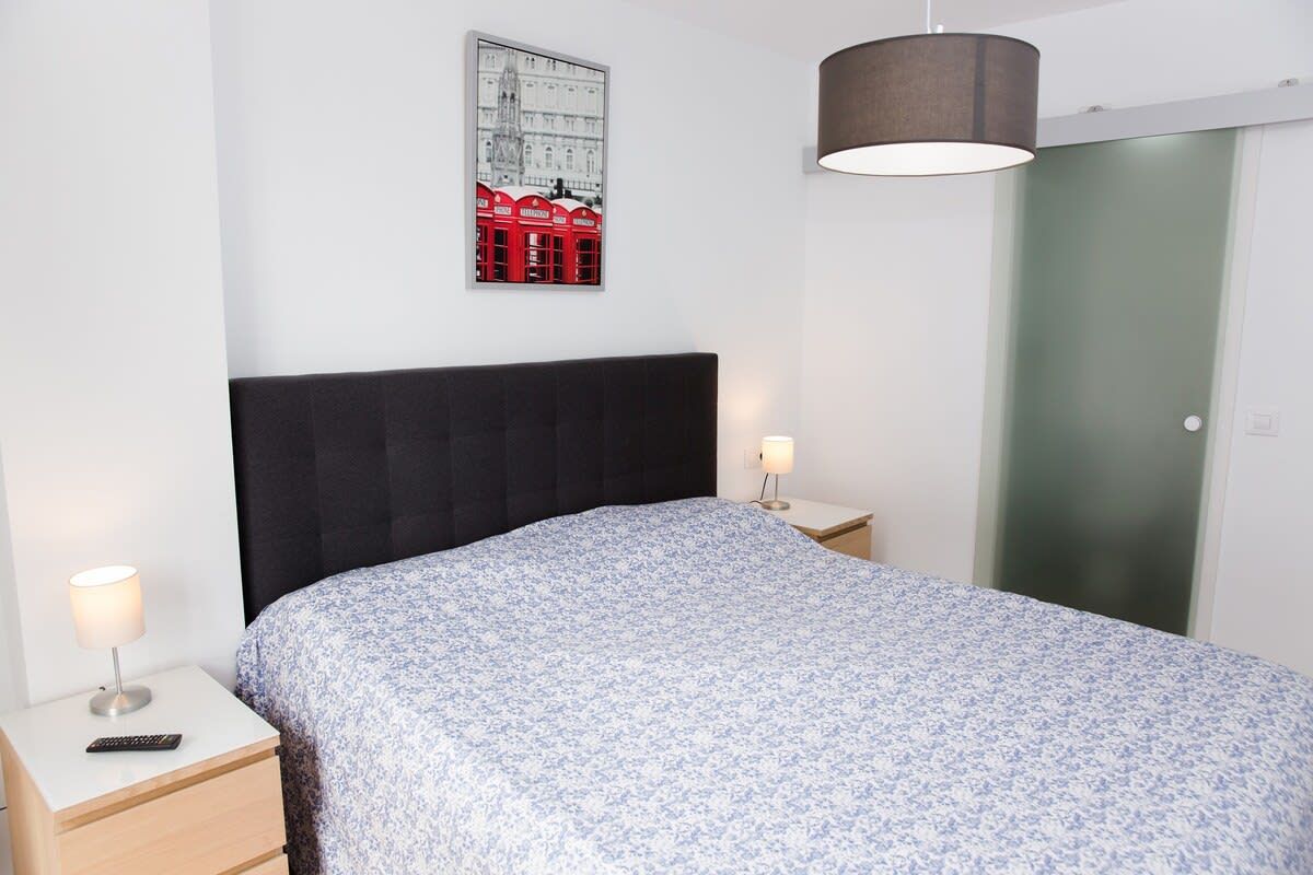 A bedroom with a bed, two bedside tables, a lamp, a framed picture on the wall, and a door.