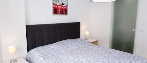 Ein Schlafzimmer mit einem Bett, zwei Nachttischen, einer Lampe, einem gerahmten Bild an der Wand und einer Tür.