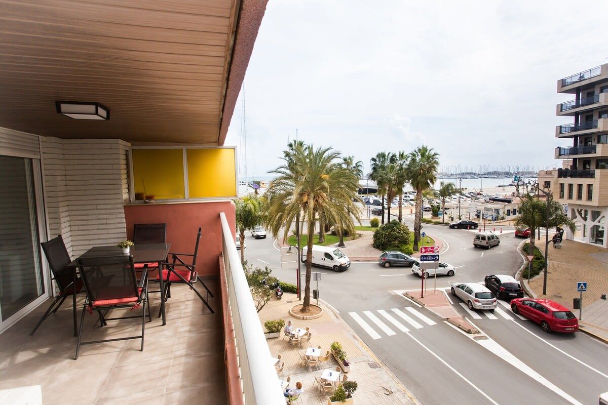 Un balcon avec une table et des chaises, donnant sur une rue bordée de voitures et de palmiers.