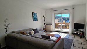 A living room with a grey sofa, a small table, a TV, and a view of a hillside through a sliding glass door. - House with view of the sea and vineyards; Near coastal path and town center (Port-Vendres)