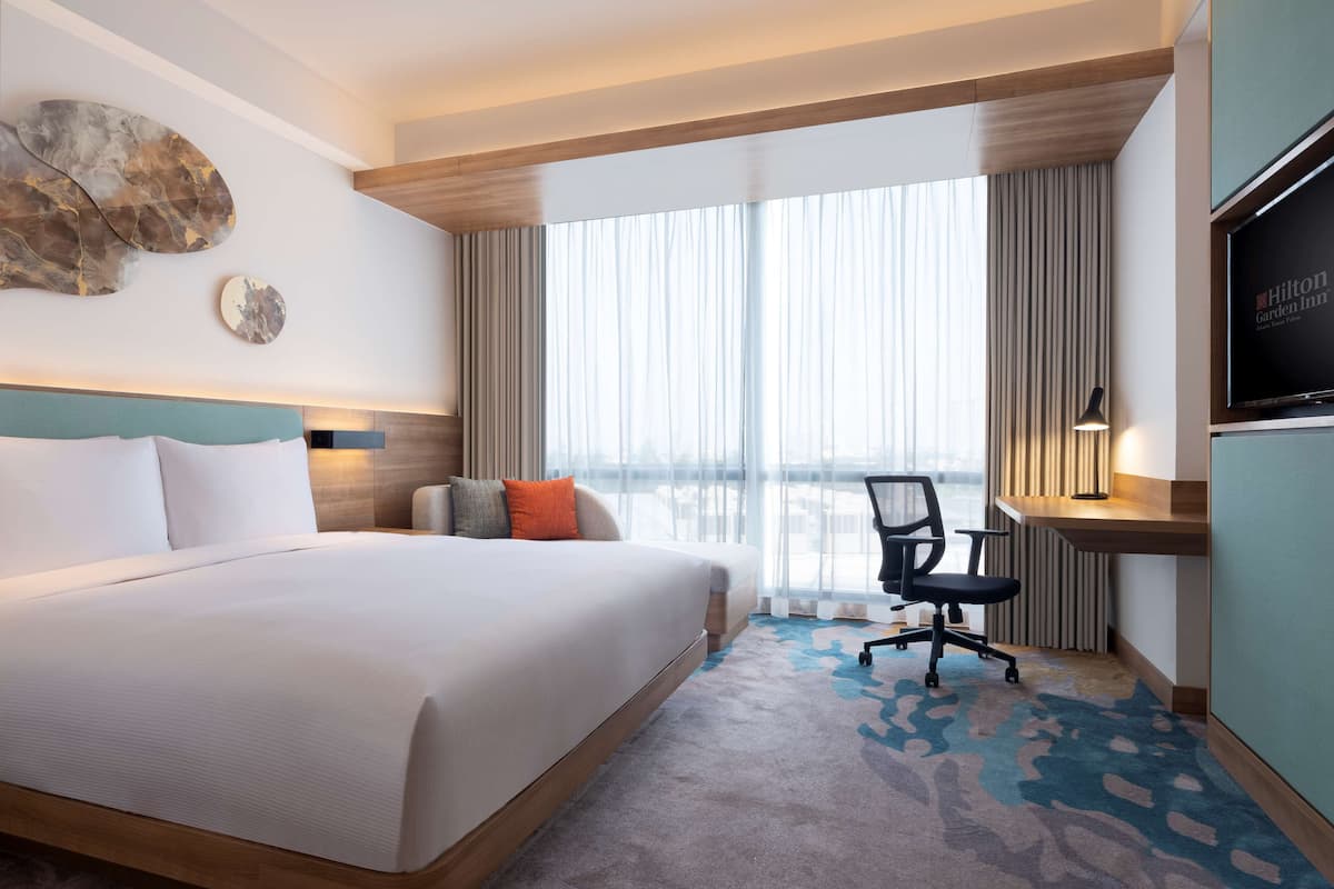 Habitación de hotel con una cama grande, un escritorio con silla, televisión y vista a la ciudad a través de amplios ventanales.