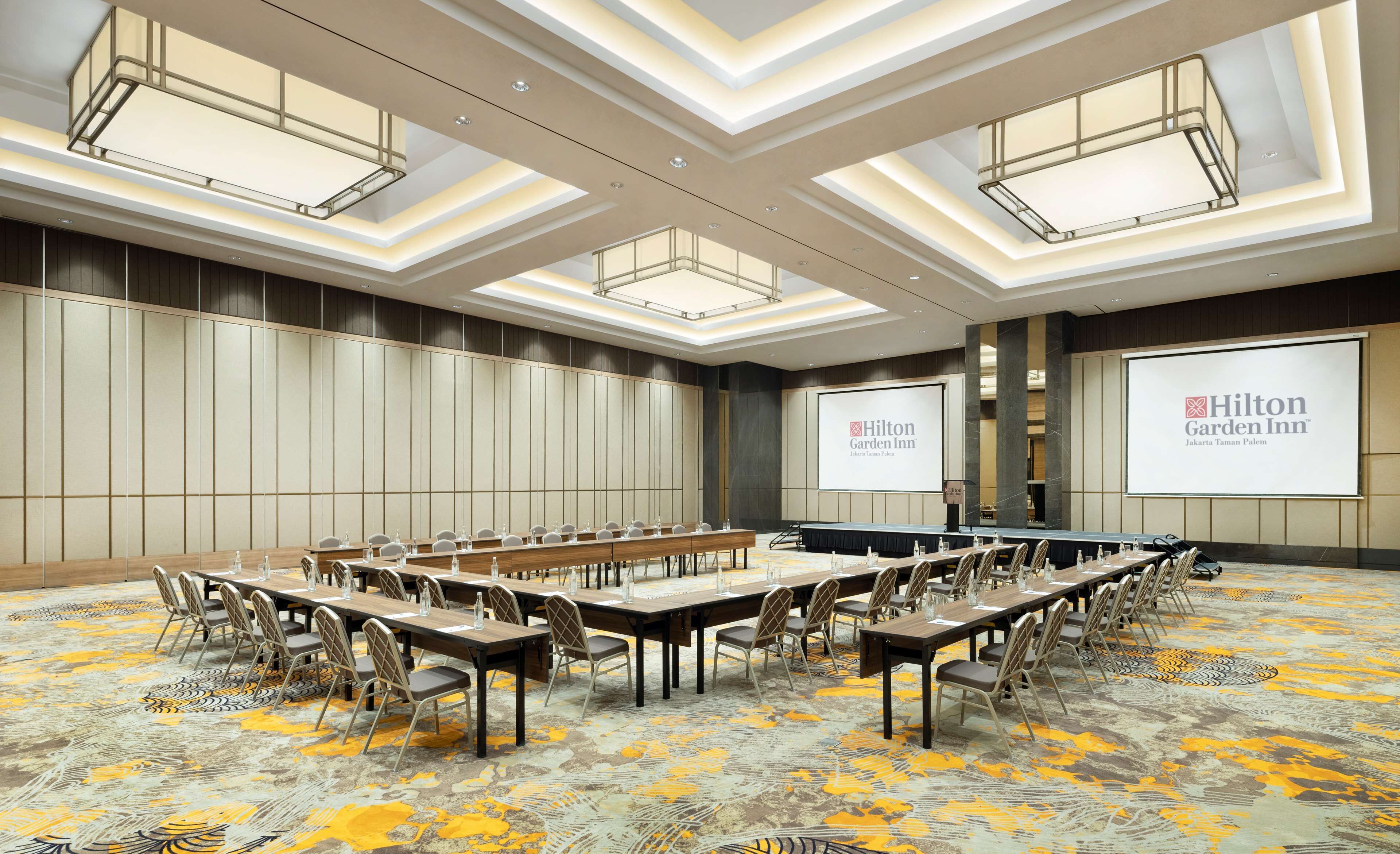 Una sala de conferències amb taules llargues i cadires, una pantalla gran que mostra el logotip de Hilton Garden Inn, i un disseny de sostre modern.