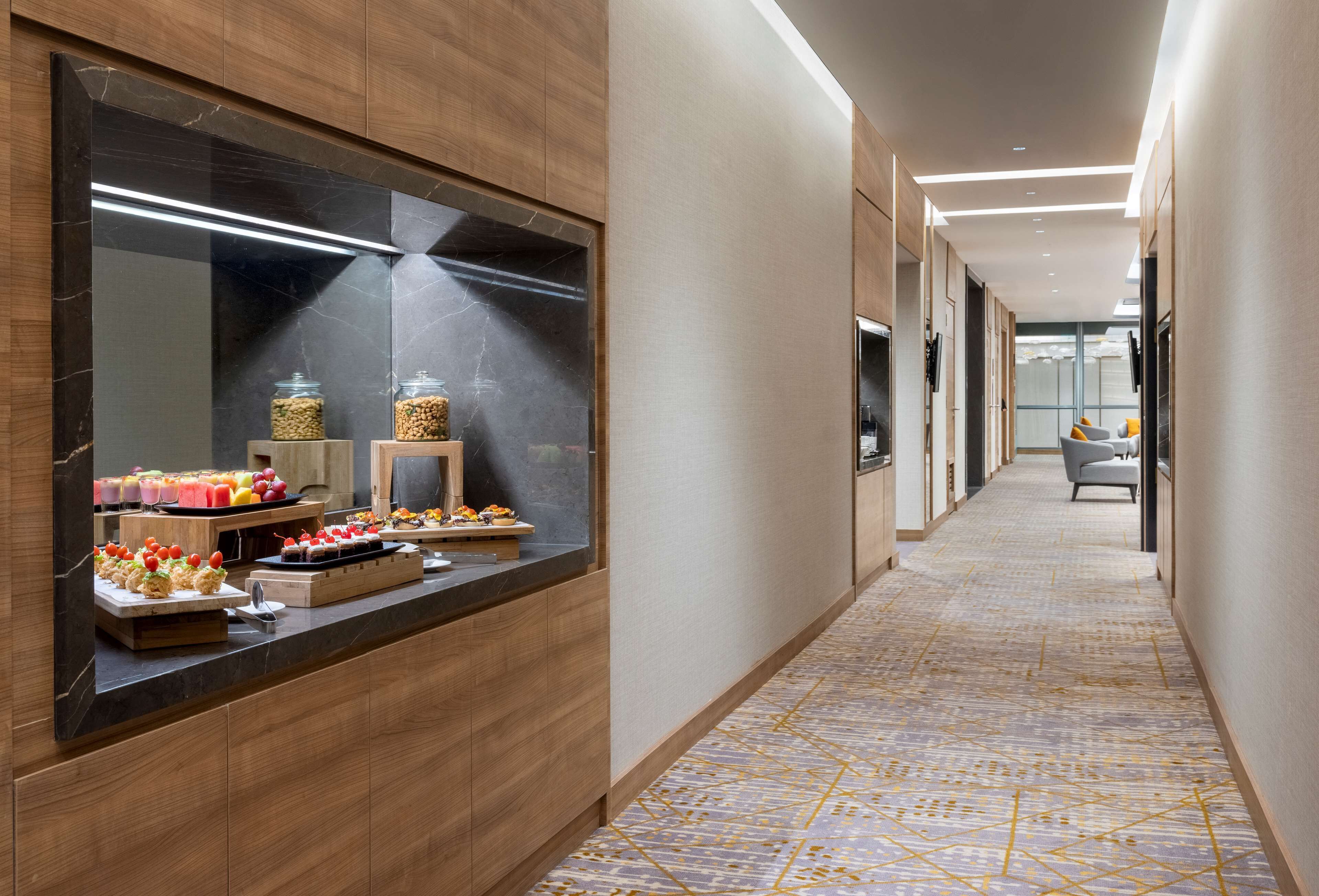 Un corridoio moderno di un hotel con un bancone in marmo che espone cibo e bevande, pannelli in legno e una moquette decorata.