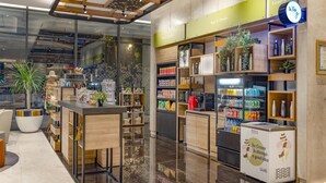 Een moderne hotellobby met een glazen scheiding, een receptiebalie en een 'Grab n Go' hoek met snacks en drankjes.