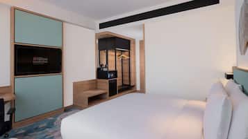 A modern hotel room with a large bed, a desk with a computer, a minibar, and a seating area with a bench.