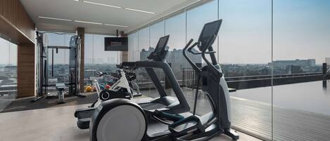 A modern gym with treadmills, stationary bikes, and weight machines, featuring large windows with a city view.
