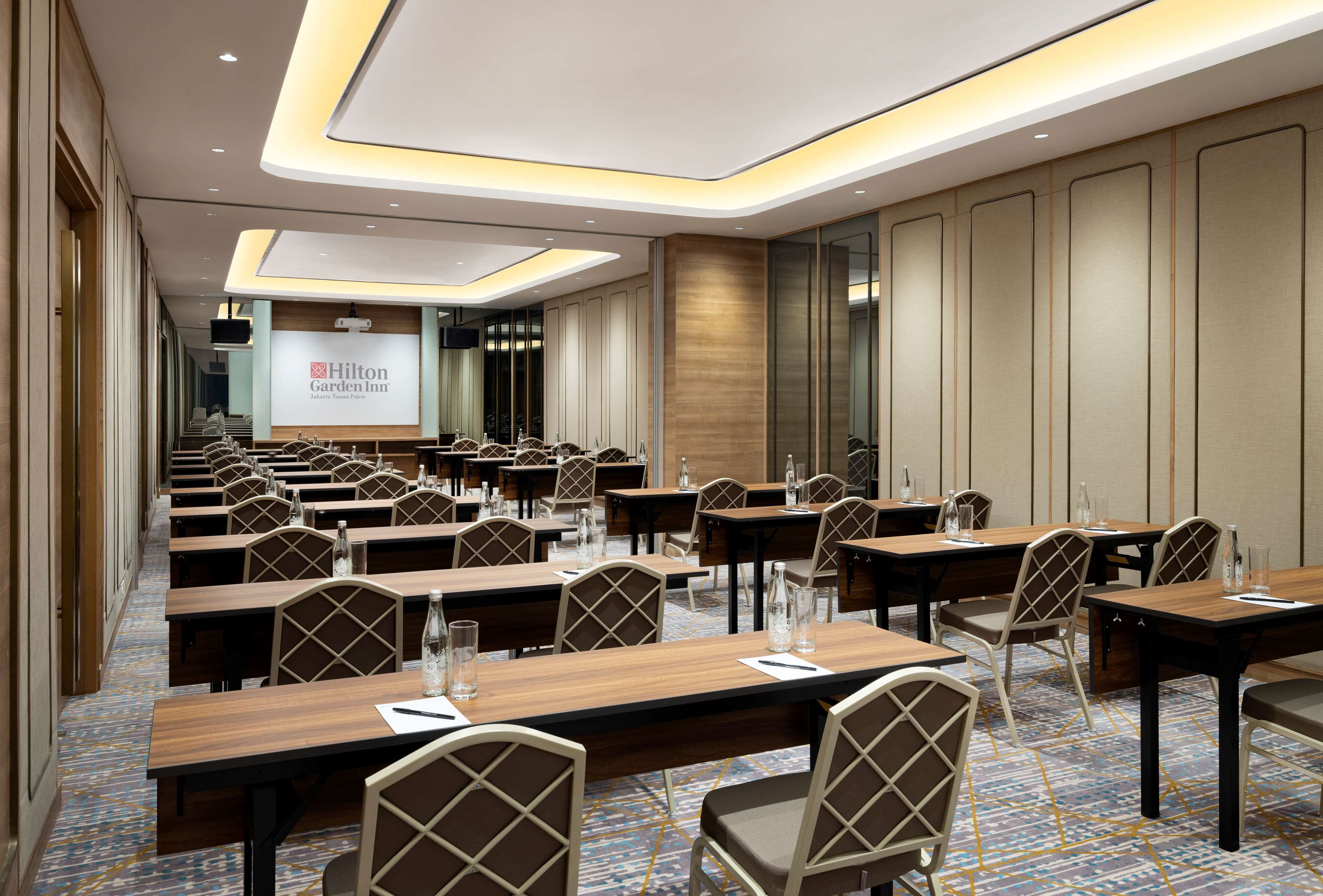 A conference room with multiple tables and chairs, a large screen displaying the Hilton Garden Inn logo, and a carpeted floor.