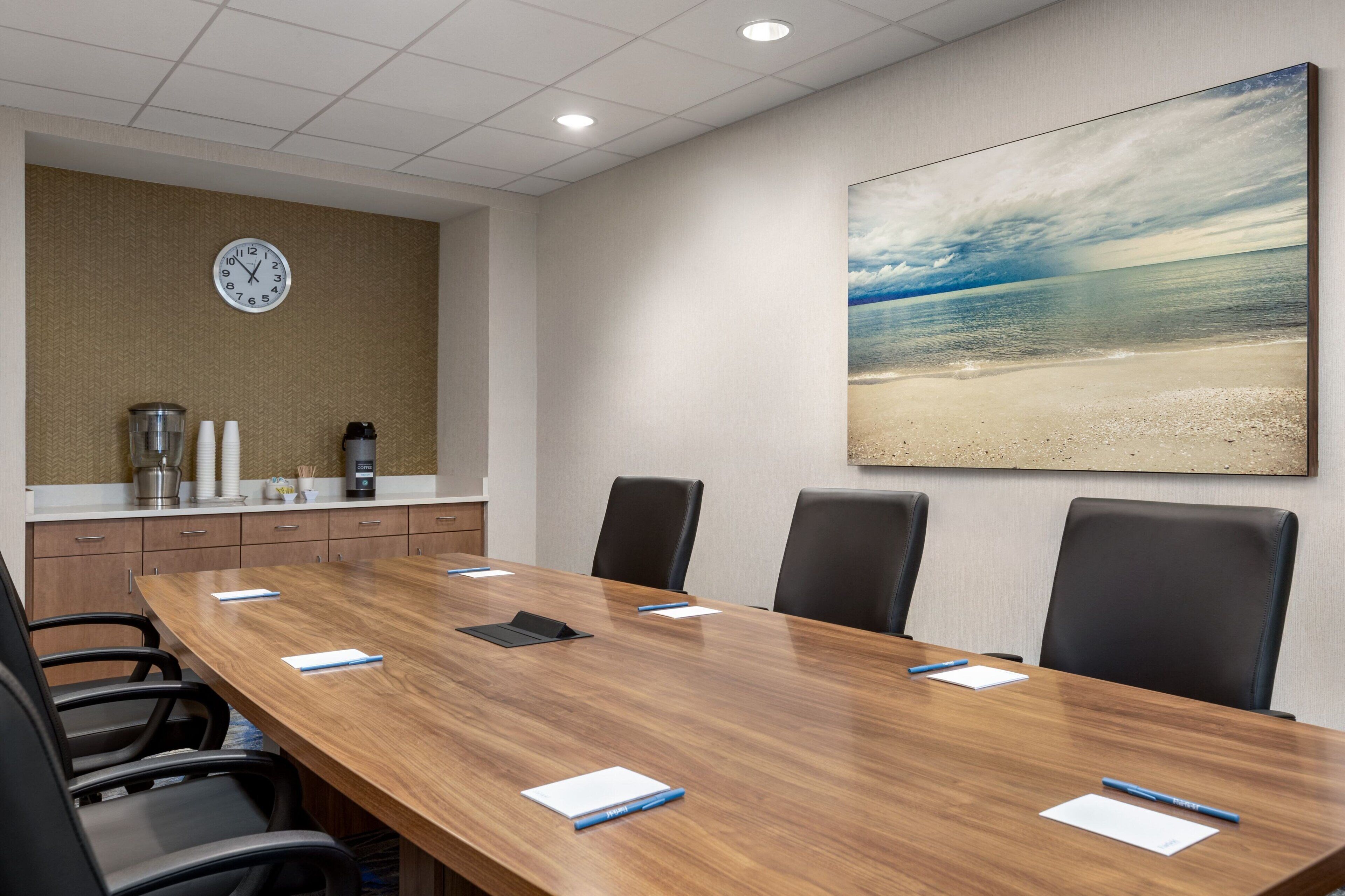 A conference room with a wooden table, black chairs, a clock, and a beach painting on the wall.