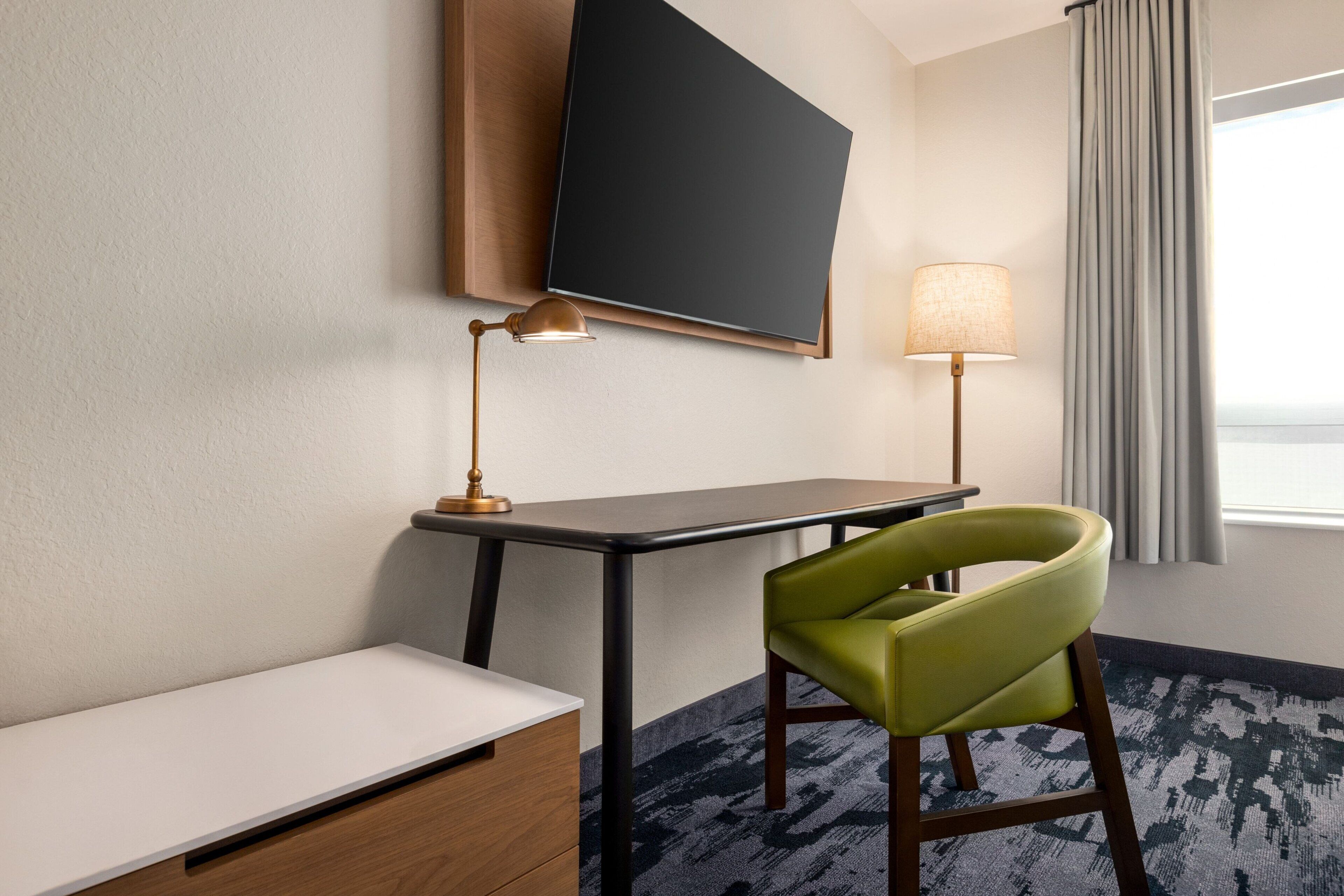 Una habitación de hotel con escritorio, una silla verde, un televisor de pantalla plana montado en la pared y una lámpara junto a la ventana.