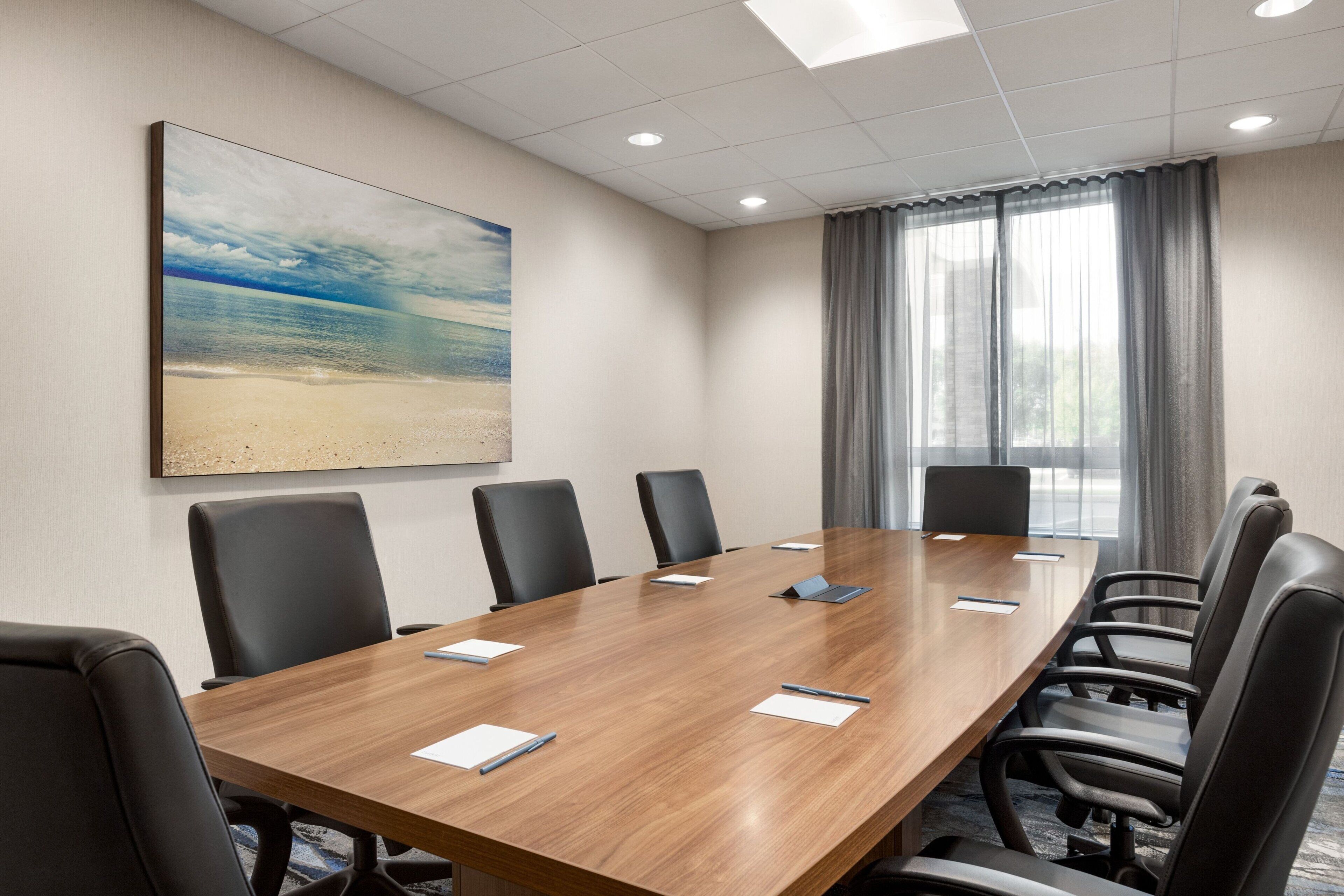 A conference room with a long wooden table, black chairs, a painting of a beach, and a window with curtains.