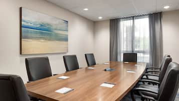 A conference room with a long wooden table, black chairs, a painting of a beach, and a window with curtains.
