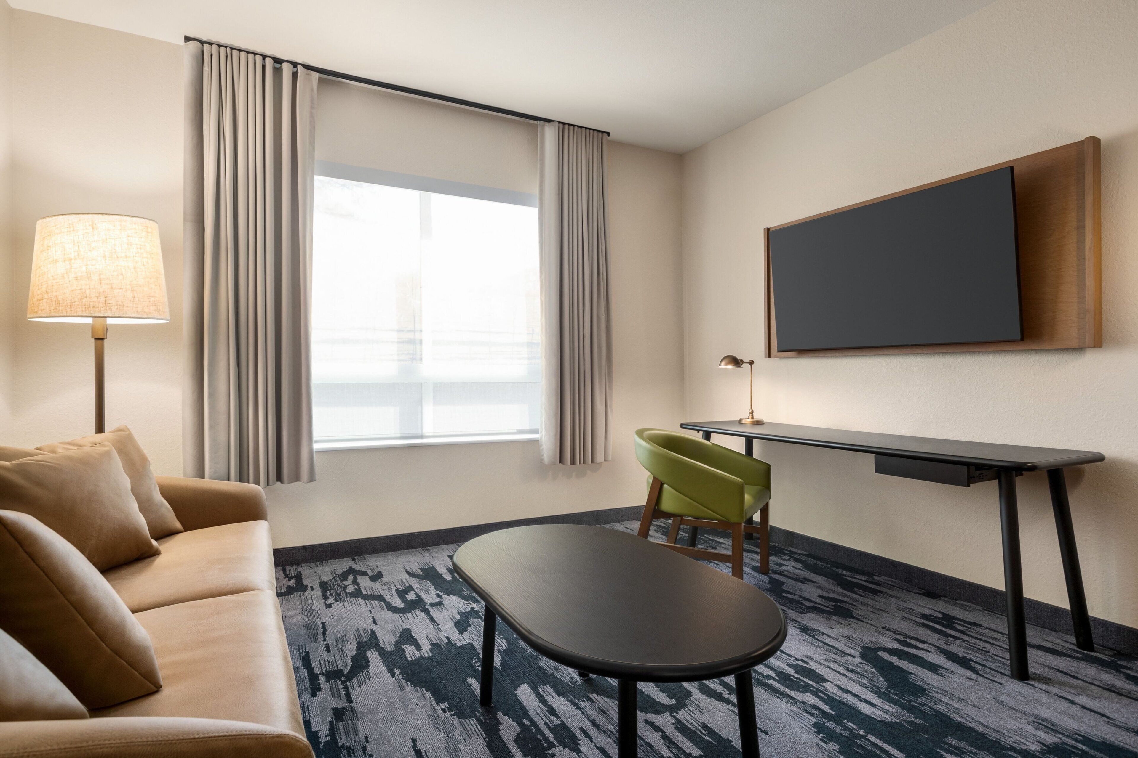 A hotel room with a sofa, a small table, a green chair, a desk, and a TV mounted on the wall.